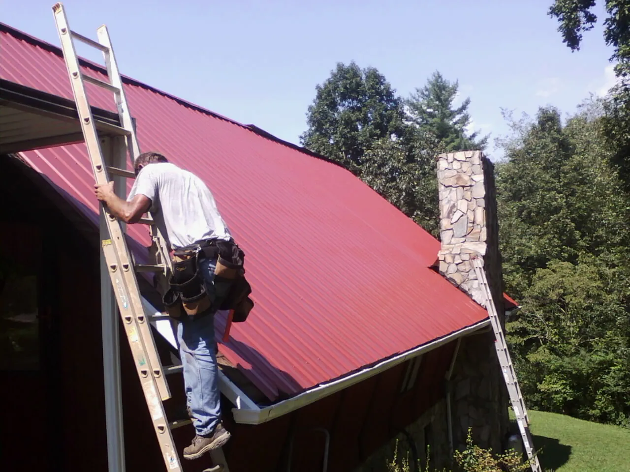 Roofer on a ladder beside a red metal roof and stone chimney — Soffit & Fascia Repair by Roofing Brothers in Homewood